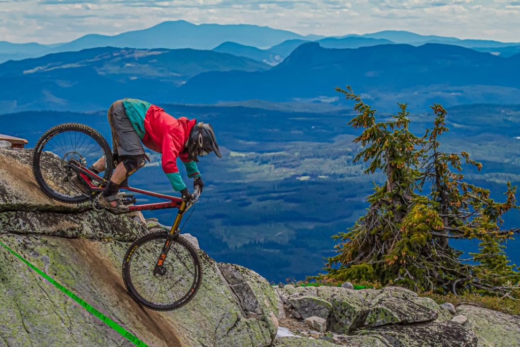 Downhill mountain bike rides down a rock feature with mountain backdrop