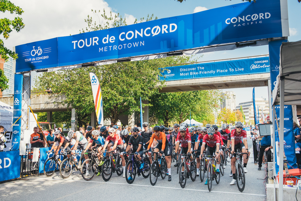 Elite Men road races line up at the start line under the Tour de Concord Metrotown banner.