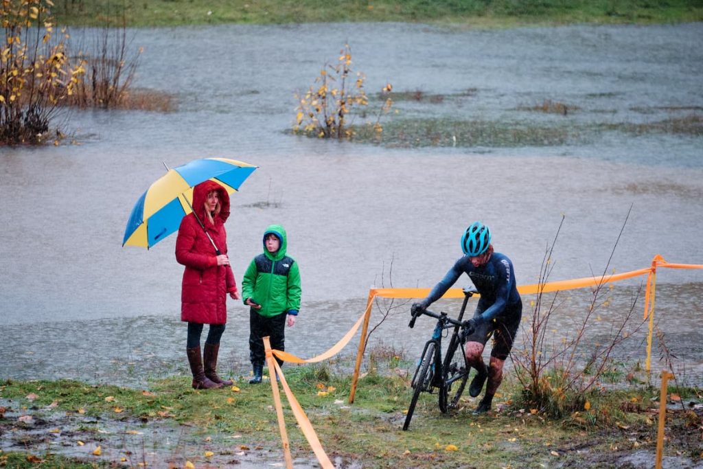 Surrounded by flooded terrain, a couple of spectators watch a cyclocross racer go by between the tape.
