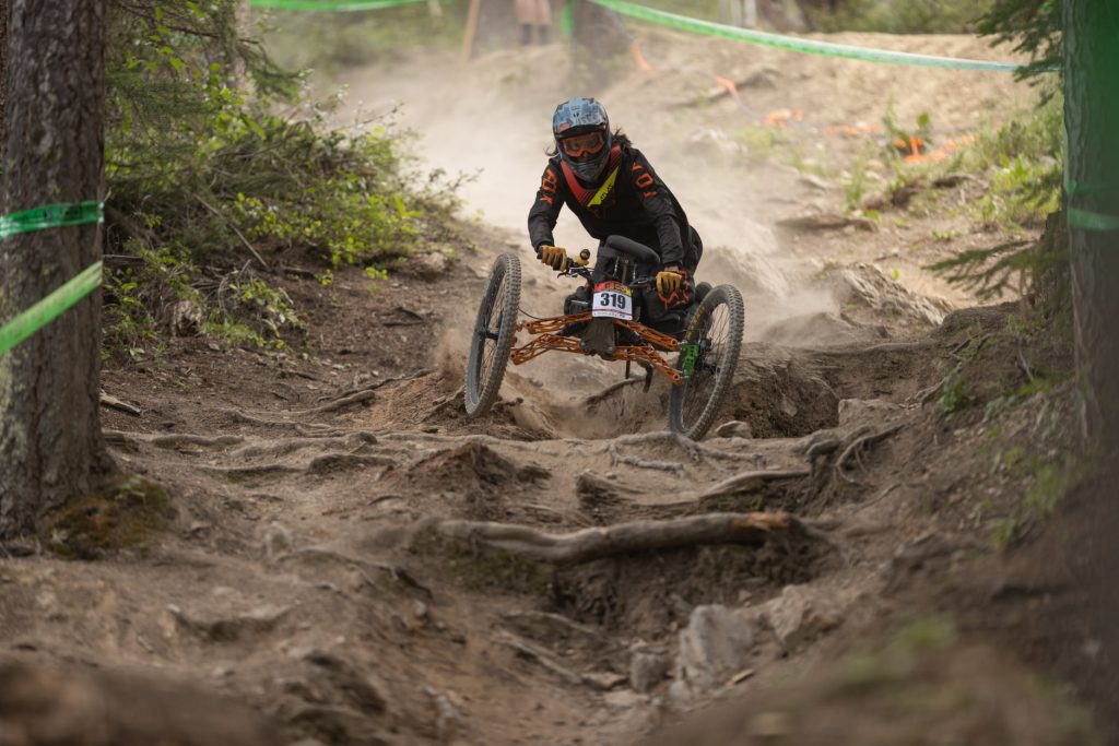 An adaptive mountain bike rider goes down a dusty run with big roots and rocks.