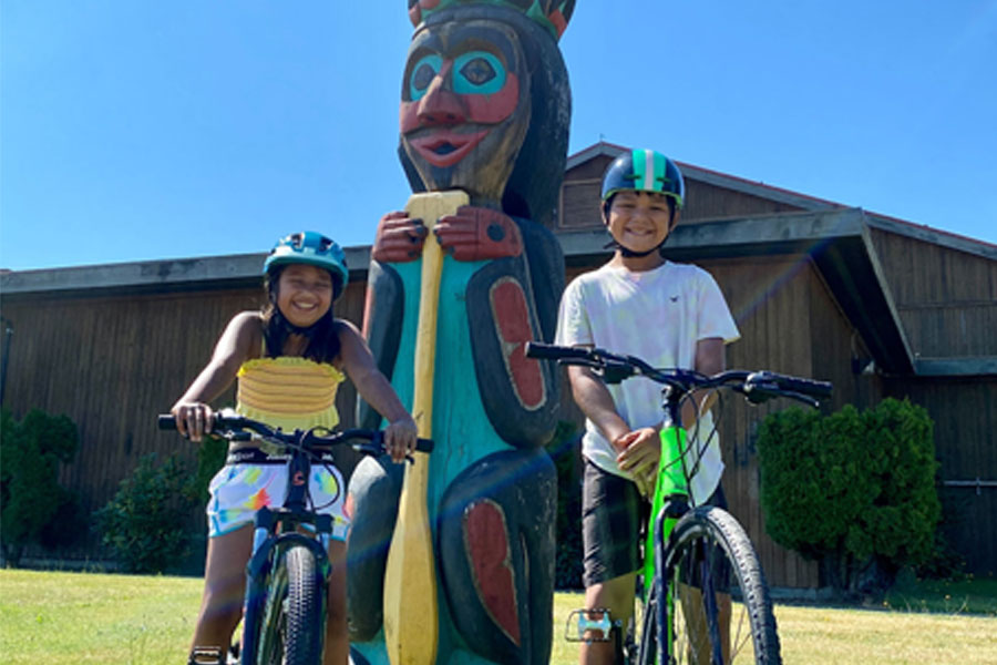 Two young cyclists smiling in front of a totem pole