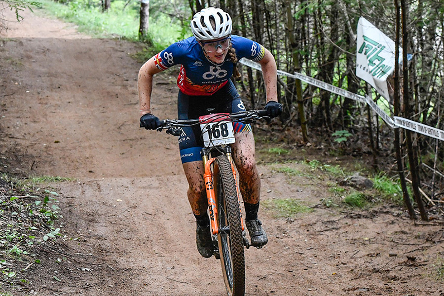 A muddied mountain biker wearing Cycling BC team kit rides along dirt path.