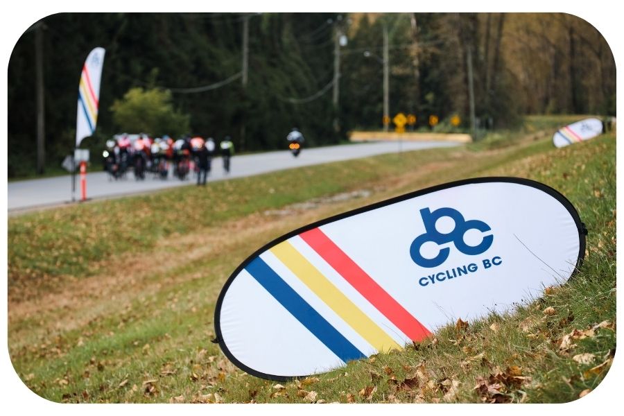 CBC sign in the foreground with the peloton riding away in the background.