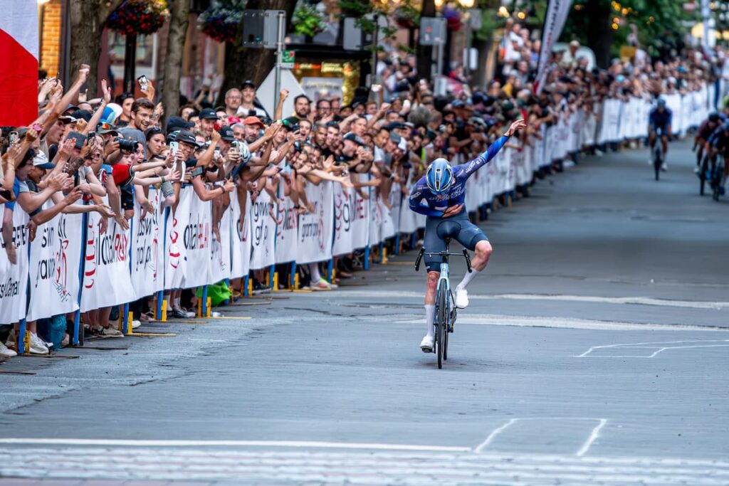 A road cyclist takes a bow while riding to the finish line while a line of fans along the side fencing cheer and take photographs.