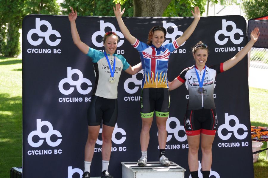 Three female athletes standing on a race podium with their arms raised.