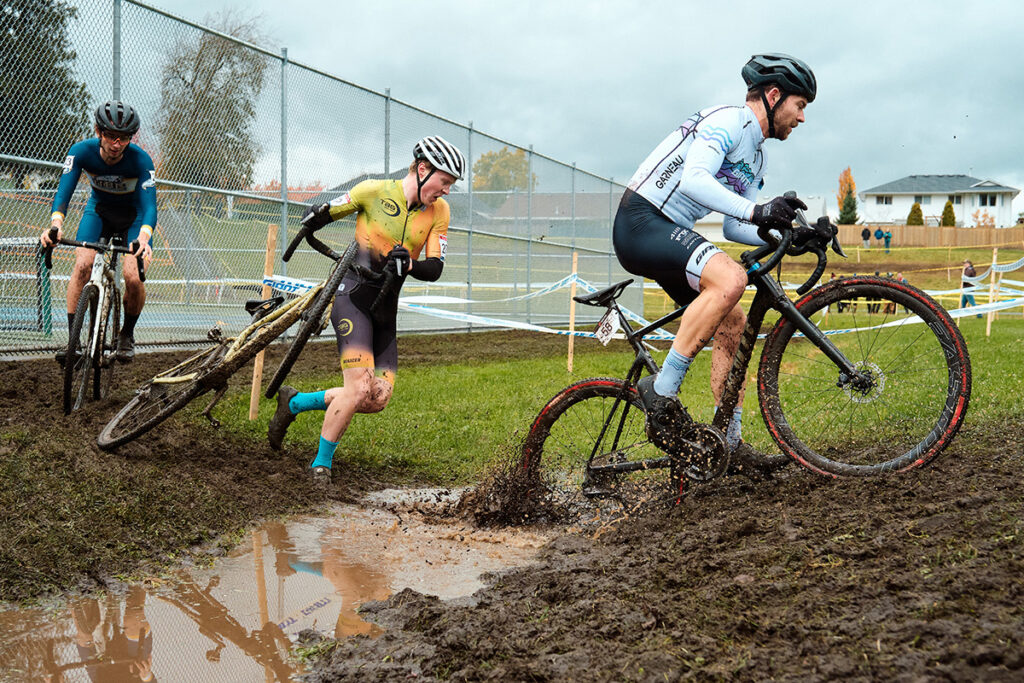 A group of three cyclo-cross racers crossing a mud puddle on their bikes and on foot.