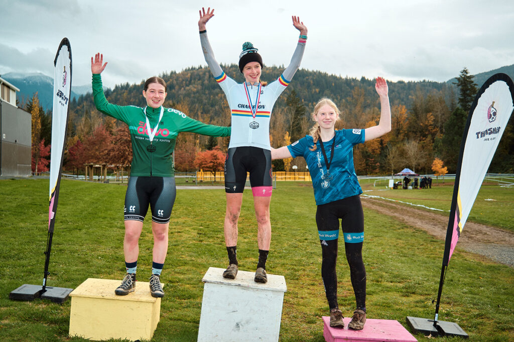 Three riders atop the awards podium on a grass field with a mountain backdrop.