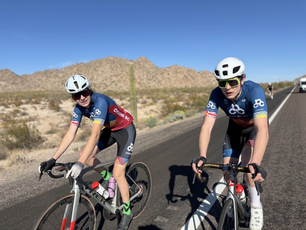 Two road cyclists in Cycling BC team jerseys on the road with a mountain range in the backdrop.