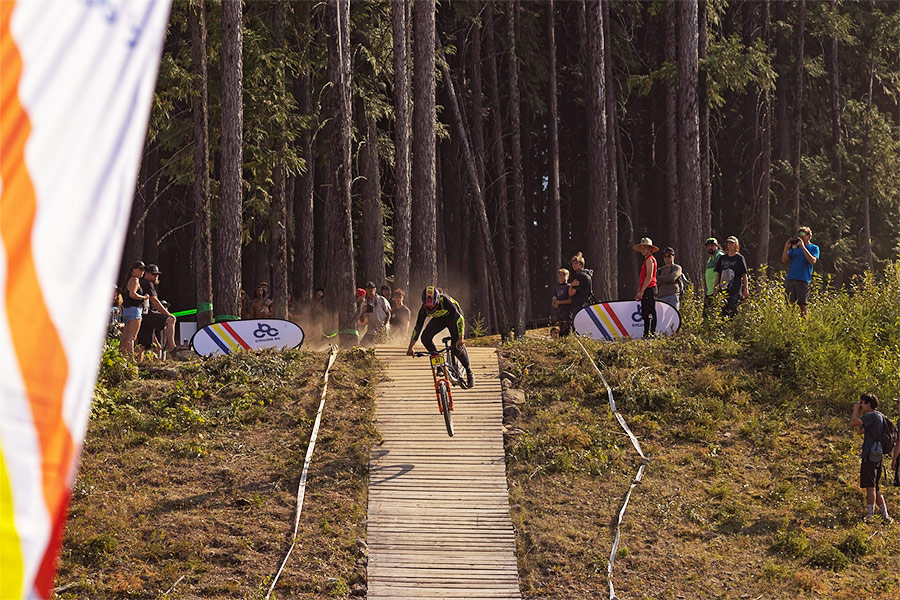 A downhill mountain biker is suspended in the air over a feature as spectators look on in the background.