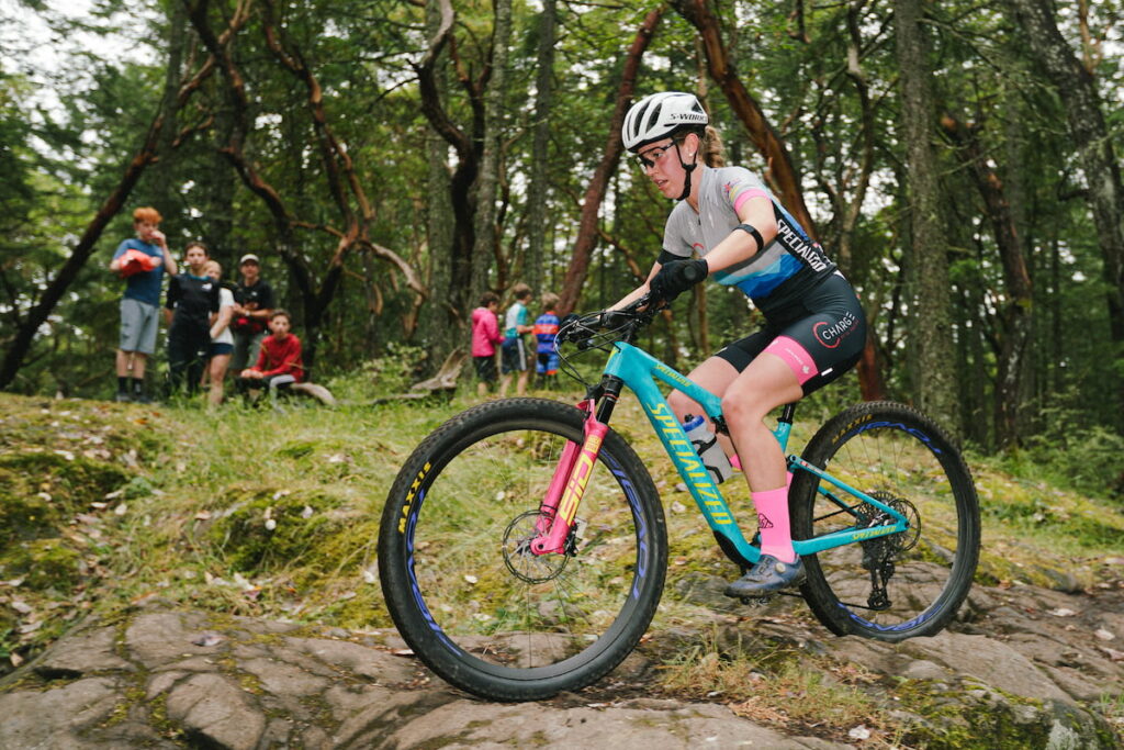 A female mountain biker going over a rock feature with onlookers in the background.