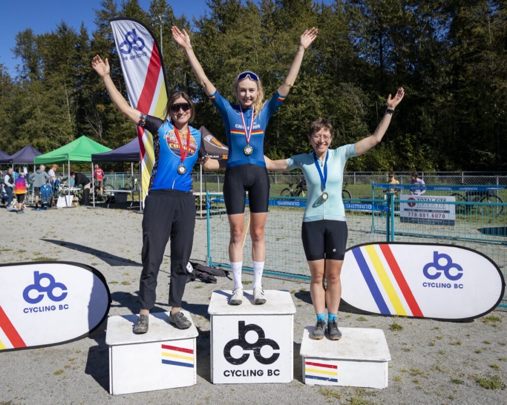 Three women adorned with medals stand on the Cycling BC awards podium on a gravel field.